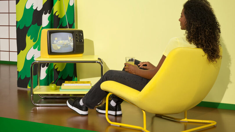 angled back view of bright yellow lounge chair with person sitting facing small yellow vintage tv on yellow cart