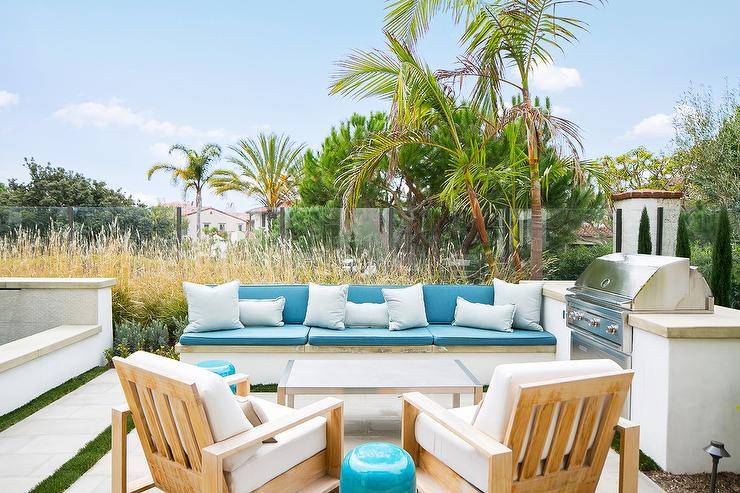 A built-in backyard bench topped with beautiful blue cushions accented with white pillows is positioned adjacent to a white outdoor kitchen finished with a stainless steel barbecue fitted to a concrete countertop. The sofa faces a stainless steel coffee table located in front of teak chairs topped with white cushions.