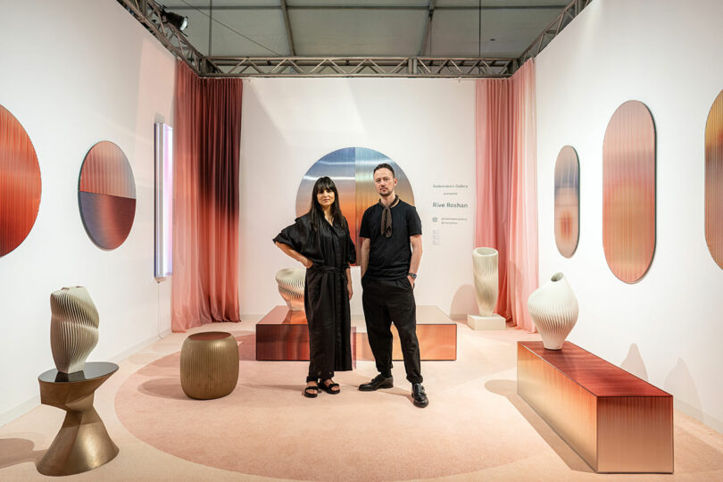 Designers standing in the center of their exhibition booth that resembles a gallery for living with home furnishings in a series orf red, pink, and nude colors.