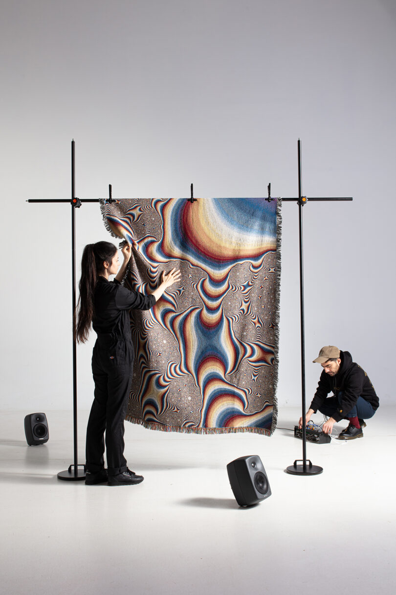 multi-colored patterned textile hanging on a rack with two speakers nearby as two people wearing all black with on it
