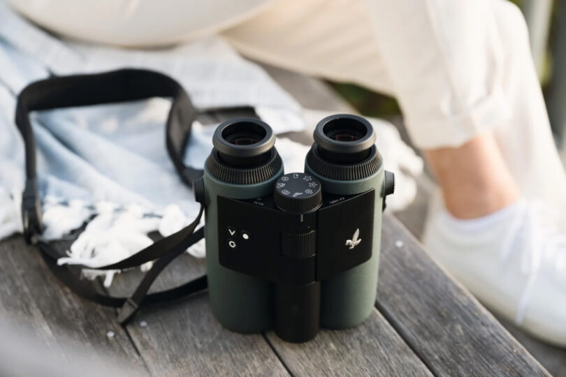 SWAROVSKI OPTIK AX Visio smart binoculars set on outdoor wooden table beside a person dress in white pants and sneakers.