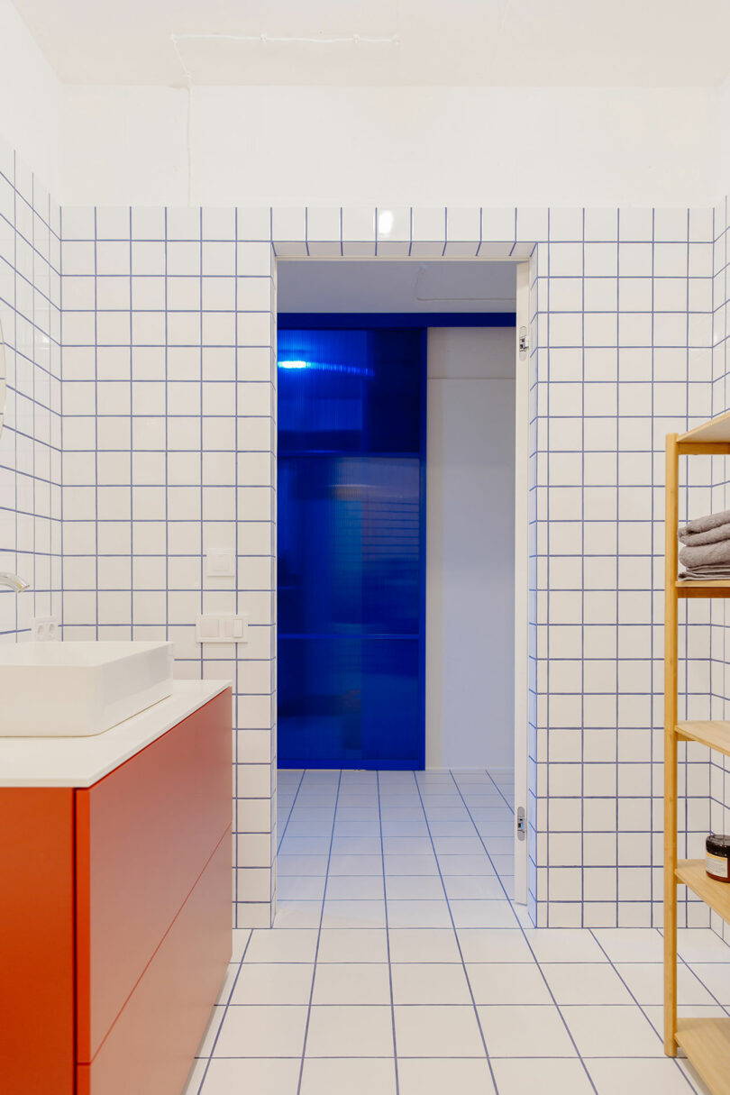 interior shot of modern bathroom with white grid tile, red sink, yellow shelf, looking into hallway