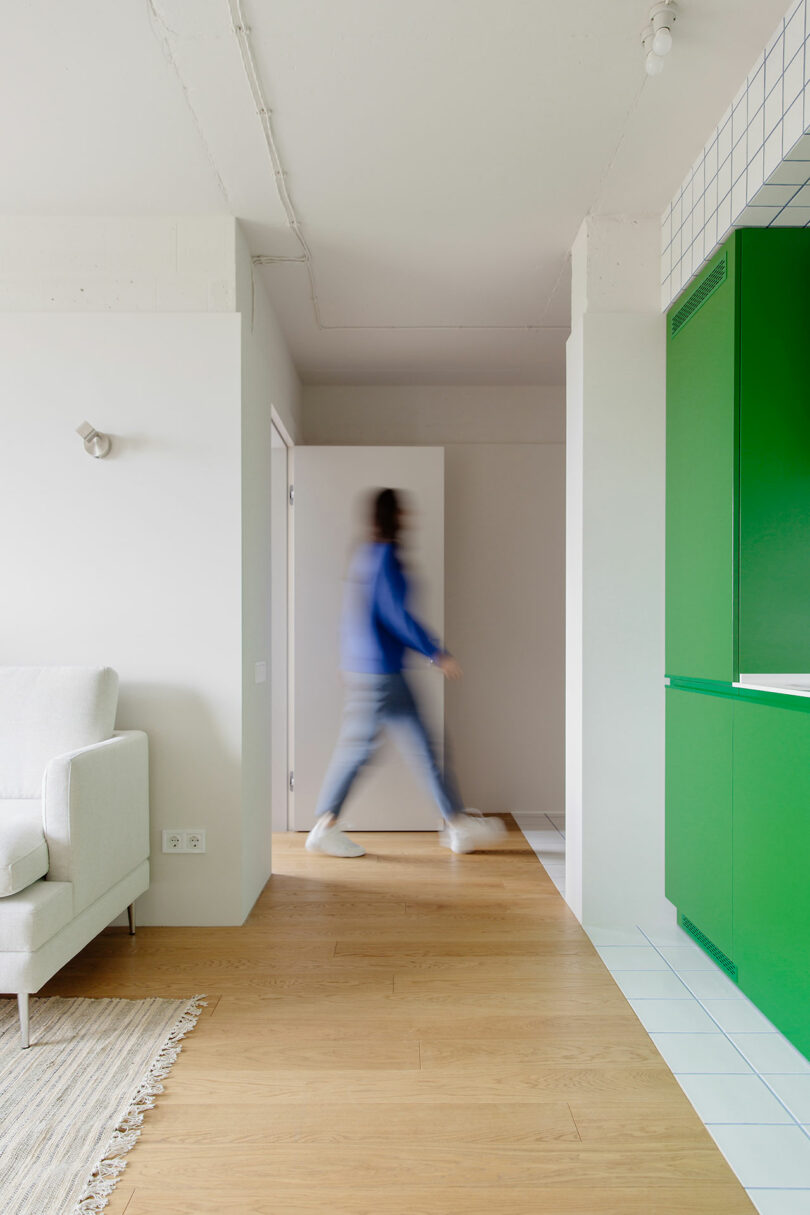 looking through modern living space with bright green kitchen on right and white sofa on left. Person in blue walking in background