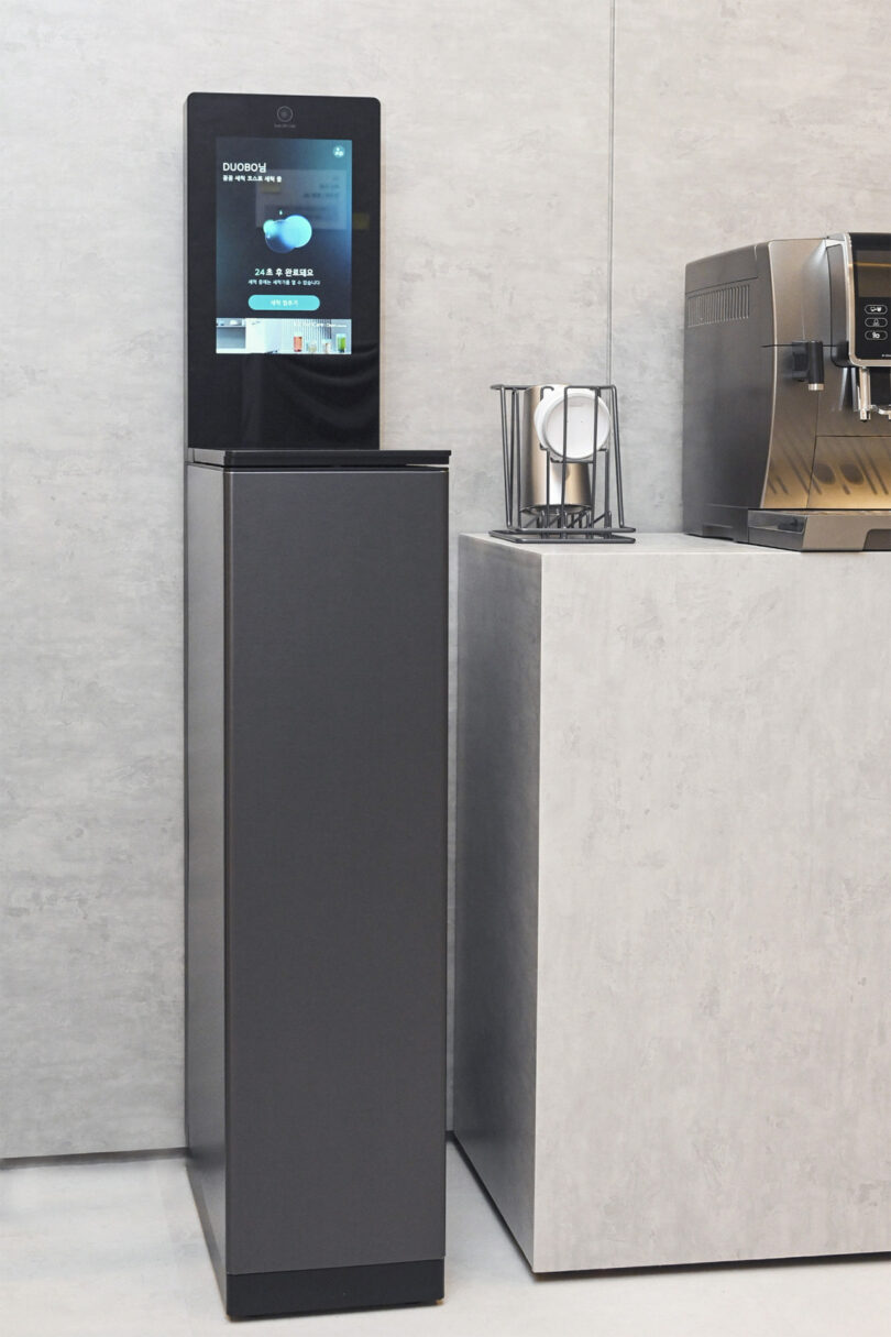 Vertically oriented single tumbler cup dishwasher with large vertical screen display stationed next to a small drying rack for a cup and a coffee machine.