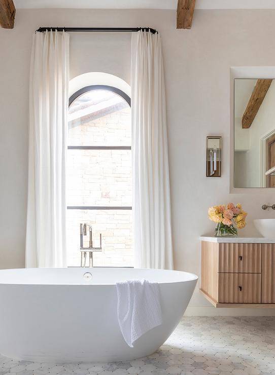 Bathroom features an oval bathtub on marble hexagon tiles under an arched window with white curtains.