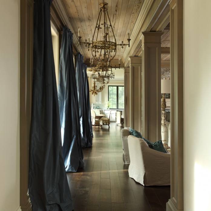 Beautiful living room features planked ceiling dotted with brass French chandeliers alongside row of windows dressed in floor to ceiling blue silk curtains.