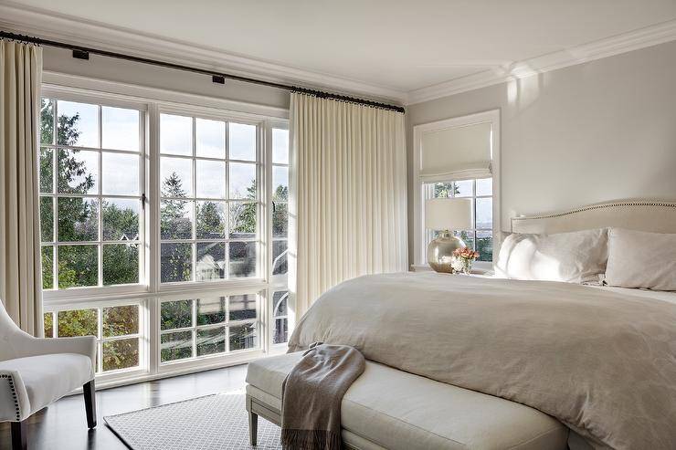 Cozy bedroom features a light tan camelback headboard lit by a mercury glass lamp, a tan bench at tthe foot of the bed and cream curtains.