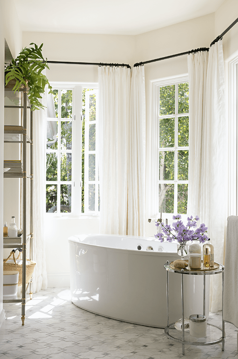 Bathroom features a bay window with oval bathtub, a round nickel and glass accent table and cream curtains.