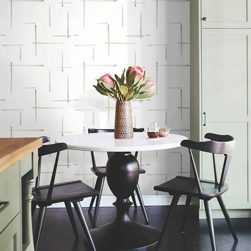 Modern dining area with a round white table, black chairs, and a vase of flowers against a wallpapered textured wall background.