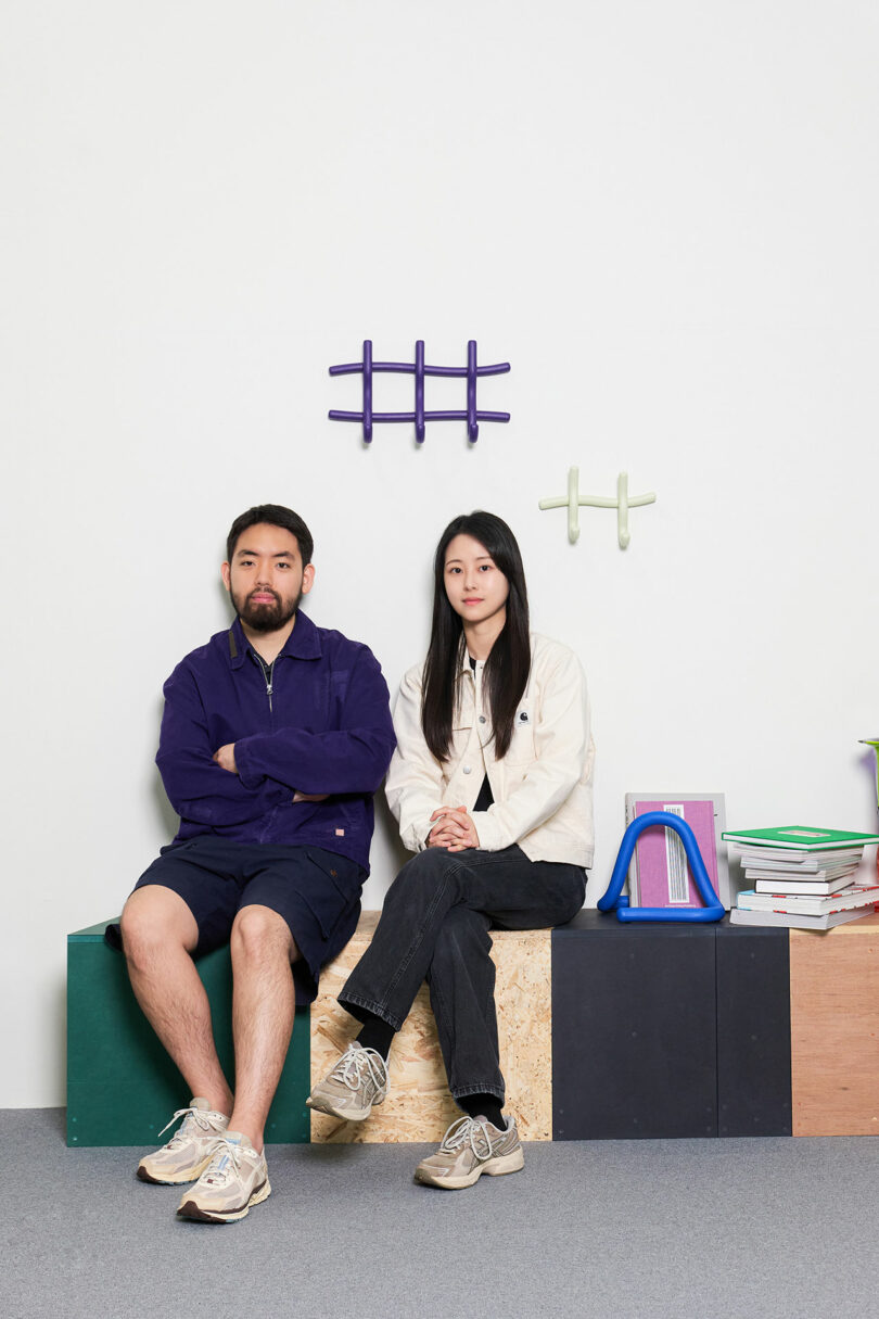 Two individuals sitting side by side with crossed legs in a room with shelves and colorful wall accents.