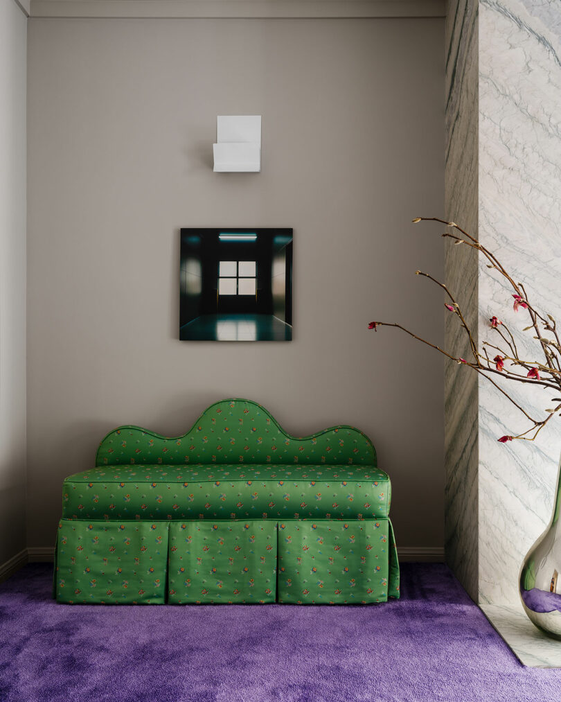 A contemporary room featuring a green patterned sofa against a gray wall, with a small painting above it, deep purple carpeting, and a vase on the floor