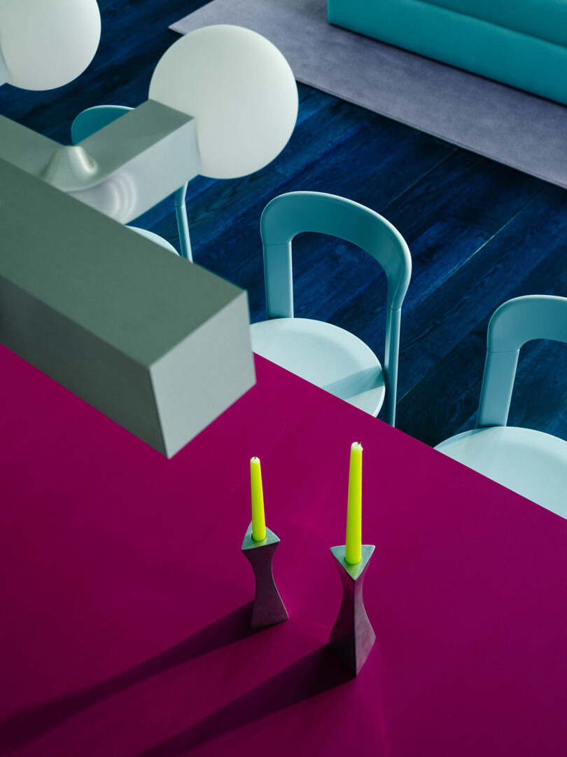 Modern dining room with a vibrant color scheme featuring a magenta table with two yellow candles, teal chairs and a blue floor.