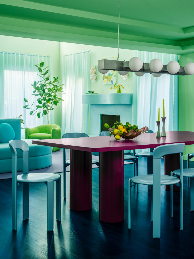 A vibrant dining room with green walls, a purple table, light blue chairs, and a fireplace with modern decor.