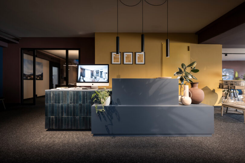 Modern office space featuring a NOA velvet-finish reception desk with adjacent yellow partition, complemented by hanging pendant lights and framed pictures.