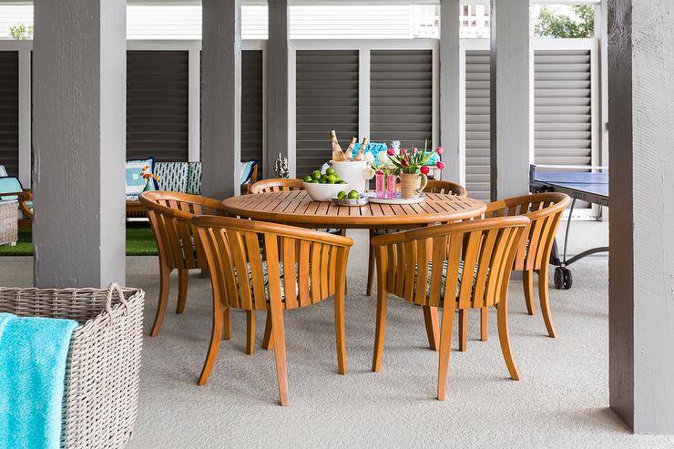 Contemporary covered patio furnished with a round teak dining table and curved teak chairs displaying a summer themed tablescape.