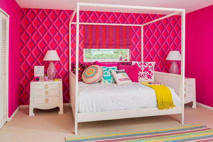 A colorful striped rug covers carpeted floors in this playful teenage girls' bedroom while positioned under a window finished with a purple and red striped roman shade framed by an accented wall covered in Pompeian wallpaper from Cole and Son's , a white canopy daybed dressed in white bedding is topped with pink and turquoise otomi pillows, an assortment of pink and accented pillows, and a mustard yellow throw. Flanking the bed are two Bungalow 5 Jacqui 3 drawer side tables topped with pink glass lamps lighting red side walls.