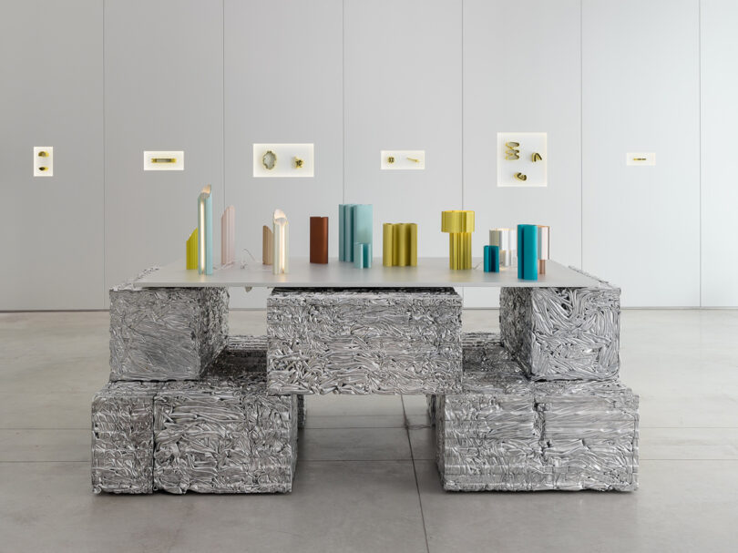 Modern art installation featuring a table and benches made from compressed metal, adorned with colorful cylindrical sculptures in a minimalist gallery space.