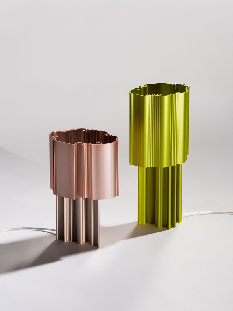 Two cylindrical, fluted table lamps in copper and green, standing on a white surface with shadows cast by ambient light.