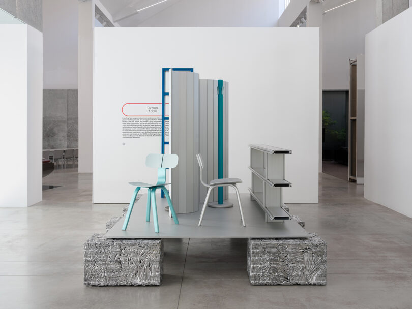 Modern art installation in a gallery featuring a minimalist chair and desk setup, structured vertical panels, and crushed metal blocks on a platform.