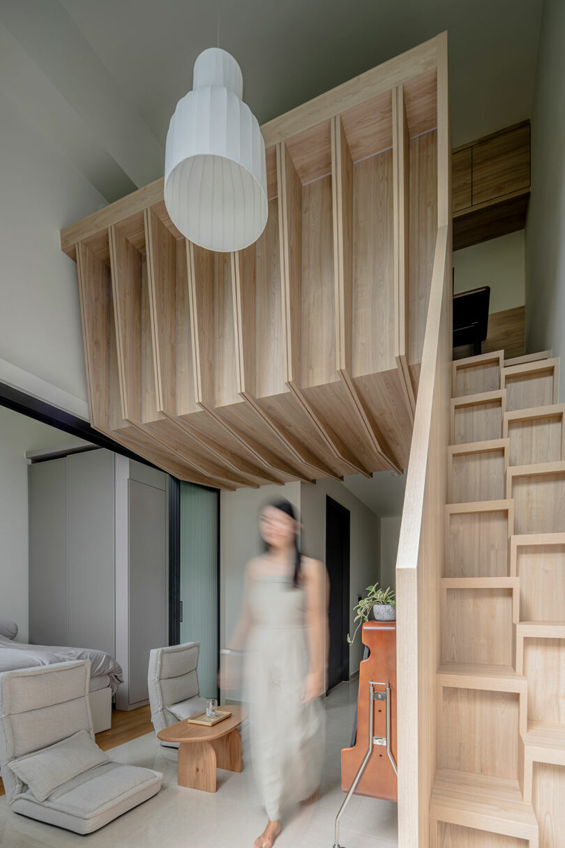 A woman walks through a modern living room with a unique wooden ceiling structure and a staircase on the right.