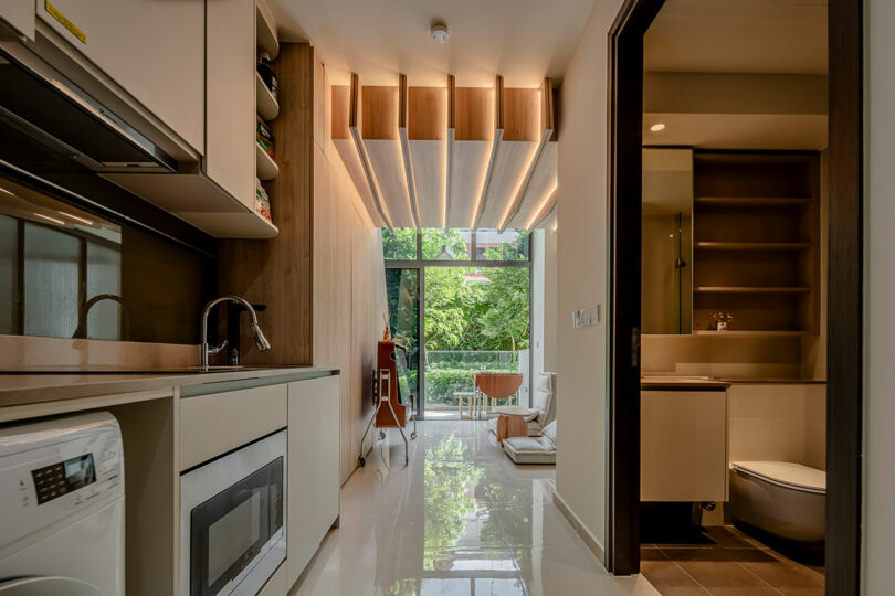 Modern kitchen with white cabinets and a washroom visible in the background, featuring stylized ceiling lights and a view of greenery outside.