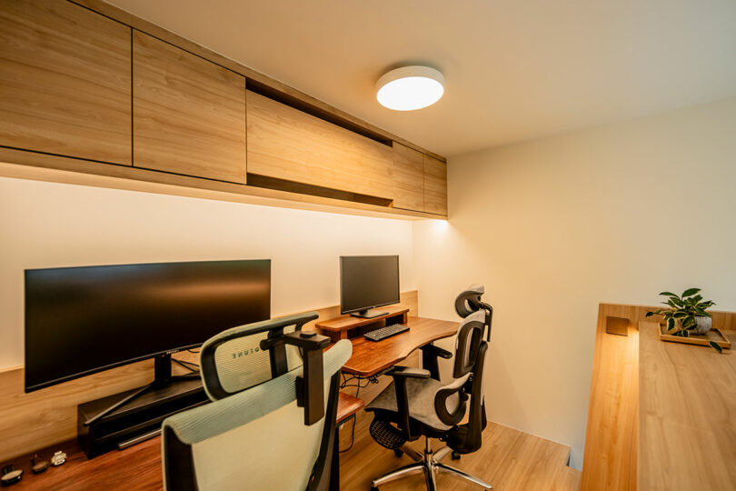 A neat home office featuring two monitors on a desk with an ergonomic chair, overhead wooden cabinets, and minimalistic decor.