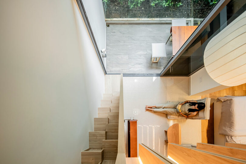 Aerial view of a person reading on a bed in a modern, compact multi-level apartment with wooden stairs and minimalistic design.