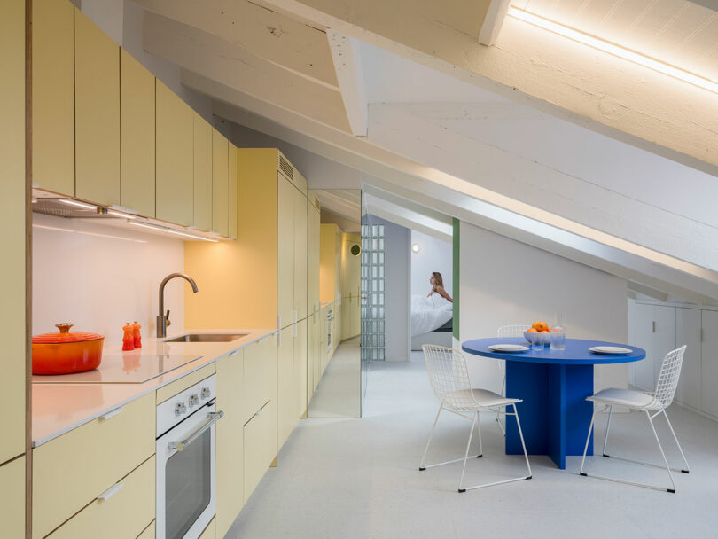 Modern kitchen interior with yellow cabinets, integrated appliances, and a dining area with a blue table.