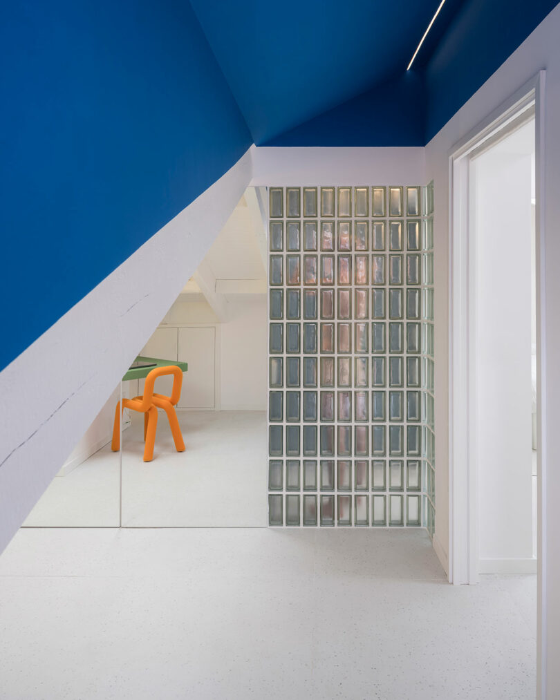 Modern attic room with blue and white walls, glass block partition, and an orange chair.