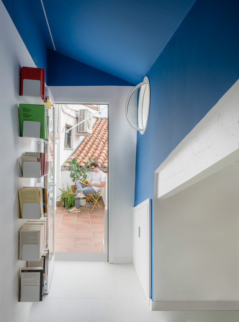 A brightly lit corridor with blue walls and ceiling, leading to a small outdoor seating area where a person is reading at a table surrounded by plants.