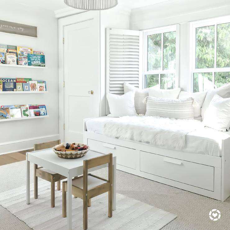 White day bed with drawers and bedding creates a restful and sophisticated appeal at a window fitted with white shutters. Layered rugs in gray and white cover wood floors in this kids play room furnished with a white play table and wooden chairs.