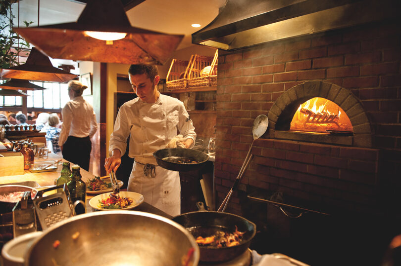 A chef cooking in a restaurant kitchen with a brick oven in the background.