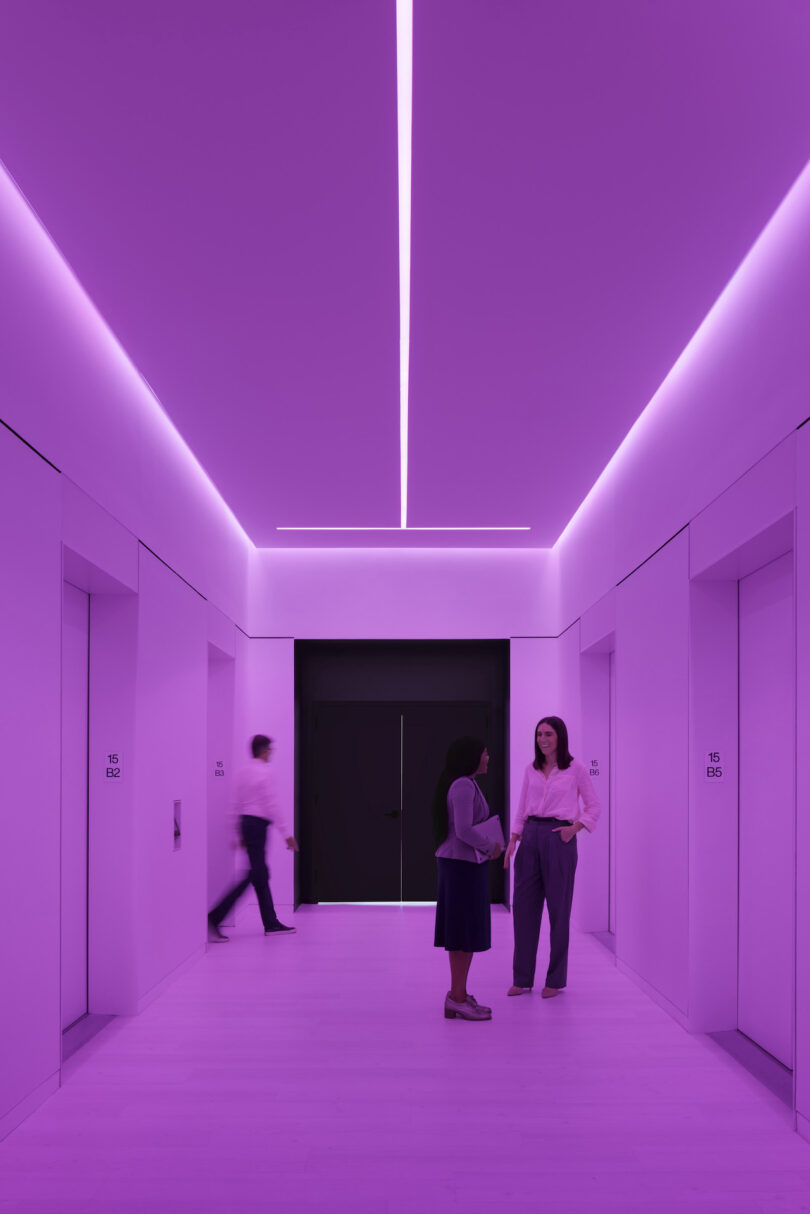 Two people converse in a hallway with vibrant purple lighting and modern design, featuring a prominent linear overhead light fixture