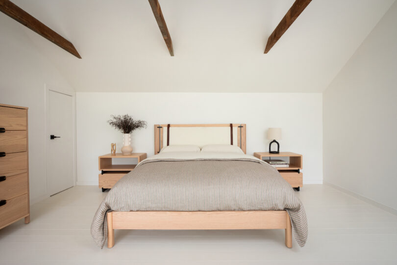 A minimalistic and contemporary bedroom with a wooden bed frame, white linens, and light wooden furniture under a pitched ceiling with exposed beams.