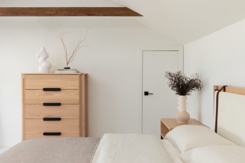 Modern minimalist bedroom with neutral tones, featuring a wooden dresser, a sculpture, and decorative vases.