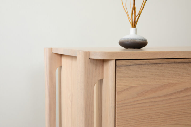 A wooden sideboard with a vase and dried twigs against a neutral background.