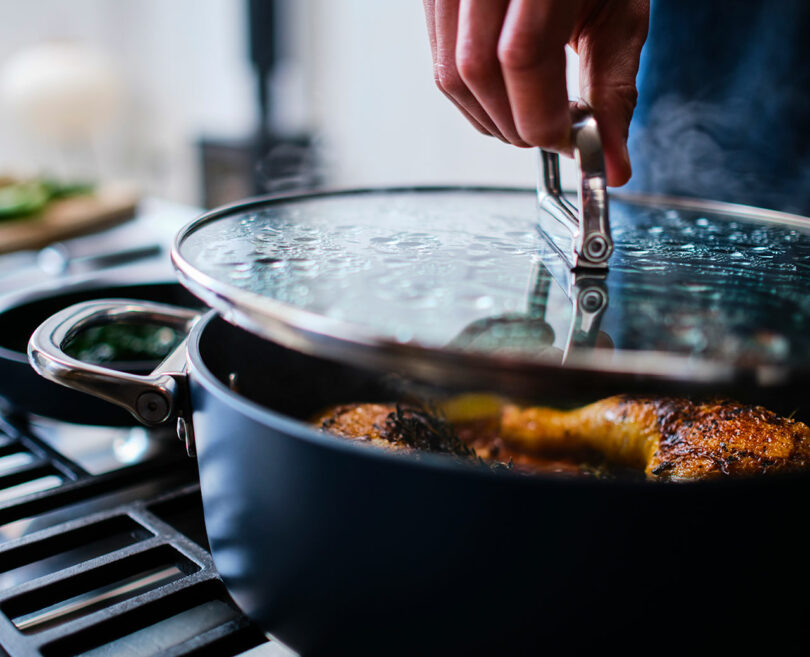 A person lifts the lid off a pan with cooking food on a stove.