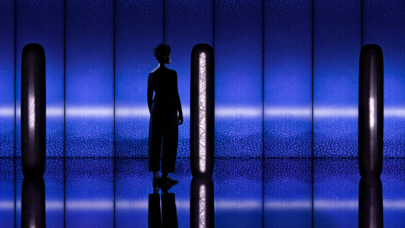 Silhouetted figure standing between glowing blue pillars in a dimly lit, textured room.