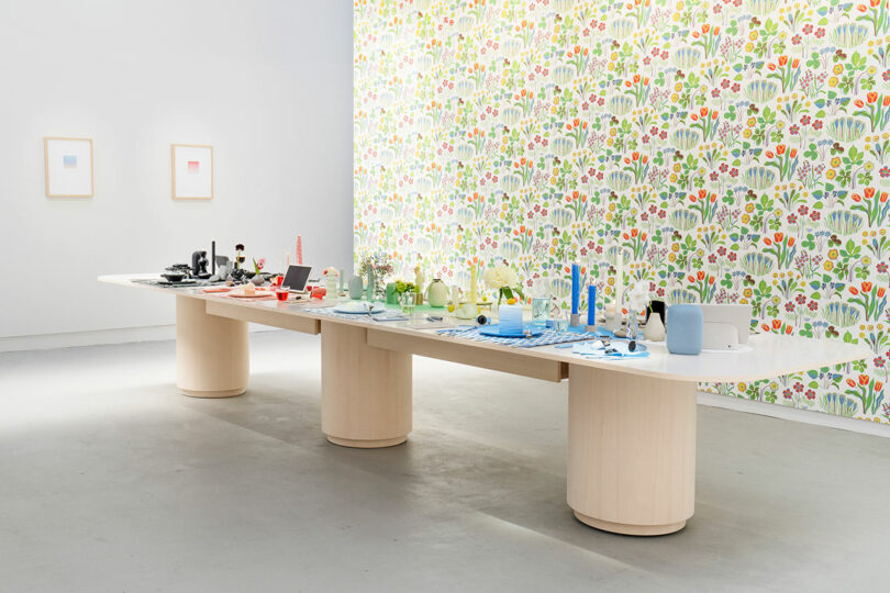 Art exhibition space featuring a long table displaying various colorful objects, flanked by floral wallpaper and framed artworks on the walls.