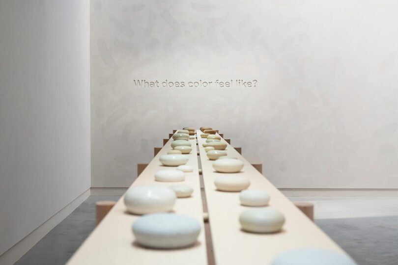 A minimalist art installation featuring rows of stones on wooden planks in a white room with the question "what does color feel like?" inscribed on the back wall.