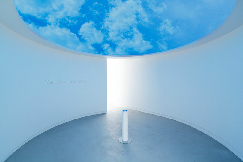 A minimalist circular room with pure white walls, a bright opening at the center, and a vibrant blue sky depicted on a domed ceiling, featuring the text "What does color look like?