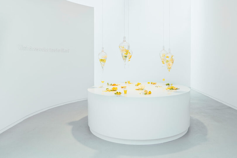 White modern gallery space with a central white circular table displaying yellow sculptures, and three hanging glass bulbs filled with yellow fragments, walls asking "what does color taste like?
