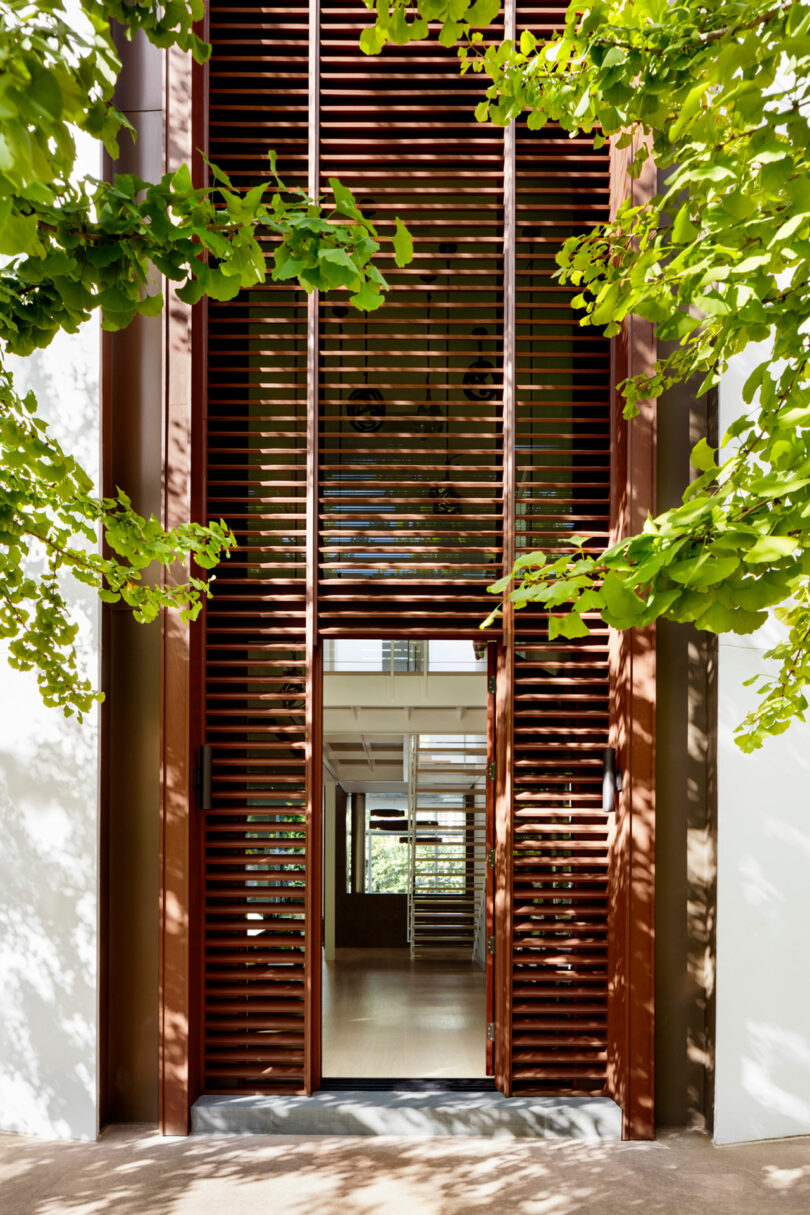 An architectural view of a modern building entrance featuring wooden slatted doors and lush greenery.