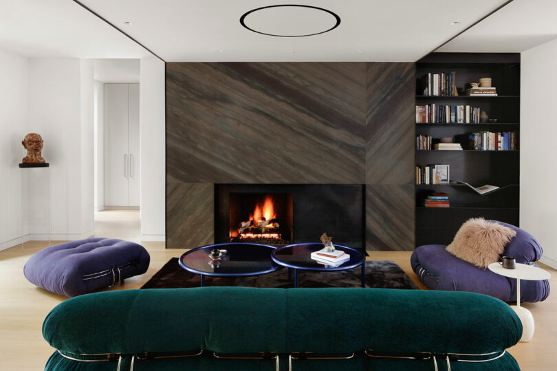 A modern living room with a herringbone-patterned fireplace surround, plush blue and green furniture, and built-in bookshelves.