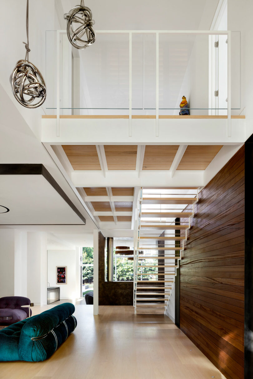 Modern interior with high ceiling, floating wooden staircase leading to an upper landing with minimalist railing, and artistic light fixtures.