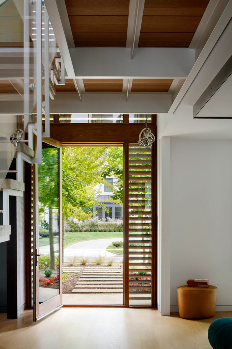 Modern home entrance with large glass door and wooden ceiling beams, opening to a garden.