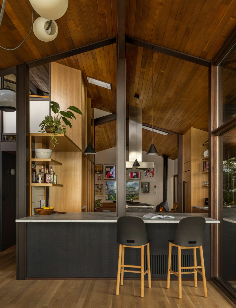 Modern kitchen with wooden ceilings and cabinets, featuring a breakfast bar with stools, and an open view into a cozy living area.