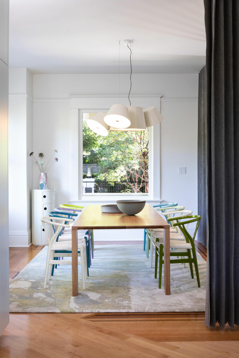 Modern dining room featuring a wooden table with colorful chairs, pendant lights, and a view of greenery through a large window.