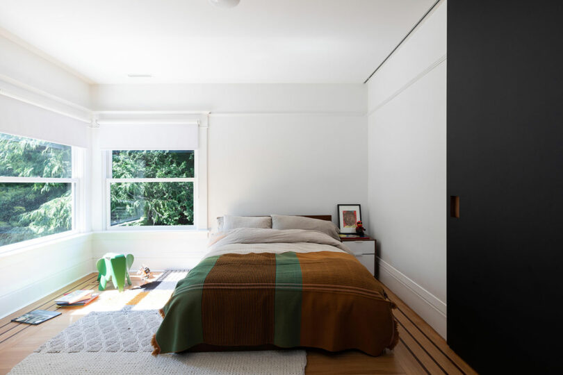 Minimalist bedroom featuring a bed covered in a green and brown blanket, a small desk with books, and a large window showing trees.