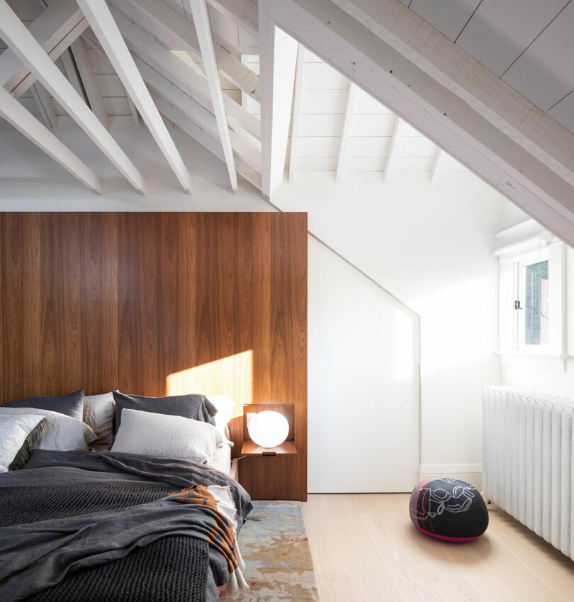 Modern bedroom design with a wooden panel headboard, white exposed beam ceiling, and a round window. Includes a bed with pillows, a lamp on a side table, and a decorative pou